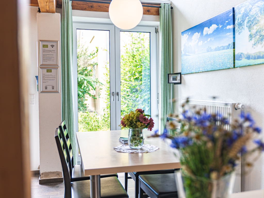Ferienwohnung 7: Esstisch mit Blumenstrauß vor einer Terrassentür mit grünem Vorhang und Blick in den Garten