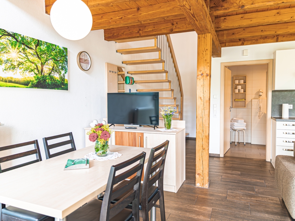 Ferienwohnung 7: Esstisch mit Stühlen und Blumenstrauß, dahinter Fernseher, Holztreppe und Blick zum Bad