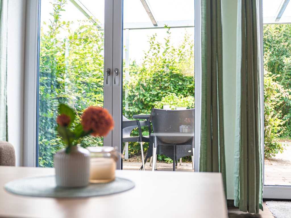 Ferienwohnung 7: Esstisch mit Blume in einer Vase, durch die Terrassentür Blick auf Stühle im Garten