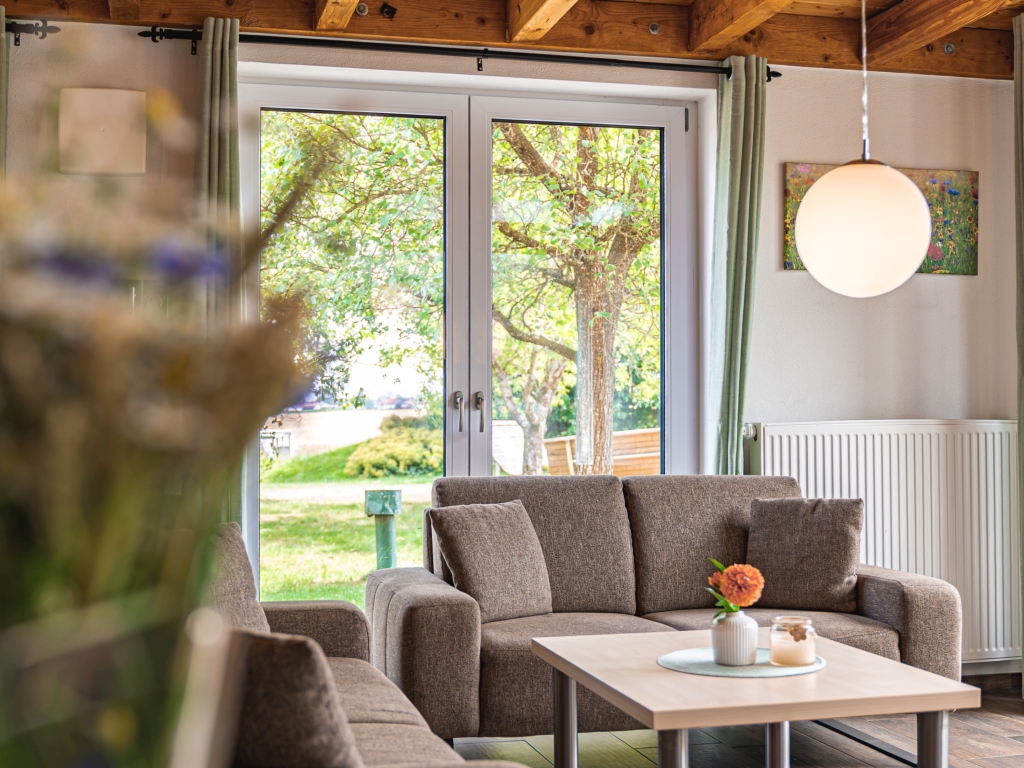 Ferienwohnung 7: Sofa mit Kissen und Couchtisch mit Blumen vor einer Fenstertür, Blick ins Grüne