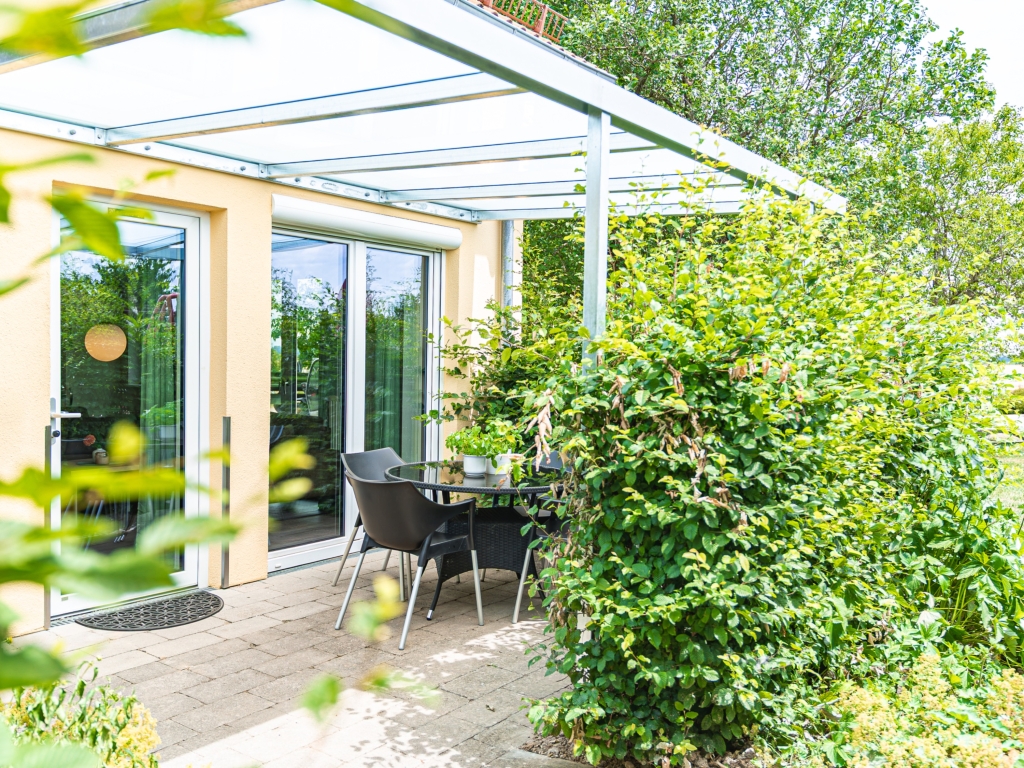 Ferienwohnung 7: Überdachte Terrasse mit Tisch und Stühlen vor der Terrassentür, daneben eine dichte Hecke