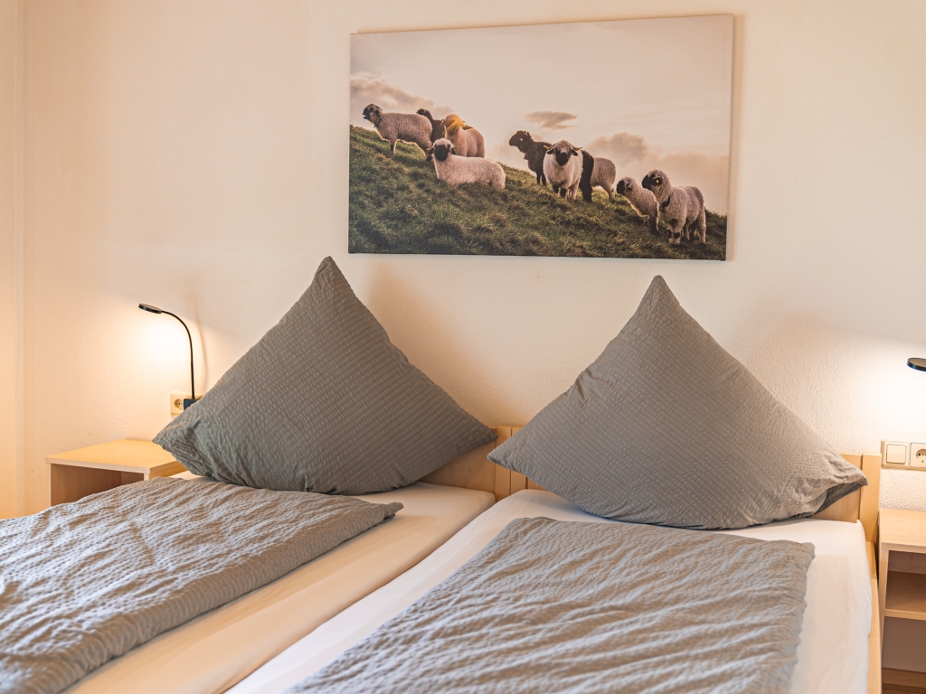 Ferienwohnung 8: Doppelbett mit grauen Kissen und Wandbild mit Tieren auf einer Weide, Nachttisch mit Lampe