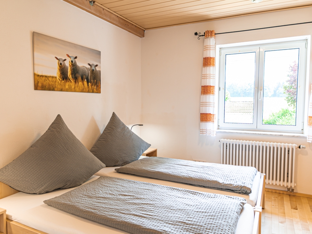 Ferienwohnung 8: Schlafzimmer mit Doppelbett, Schafbild an der Wand und Fenster mit orange gestreiftem Vorhang