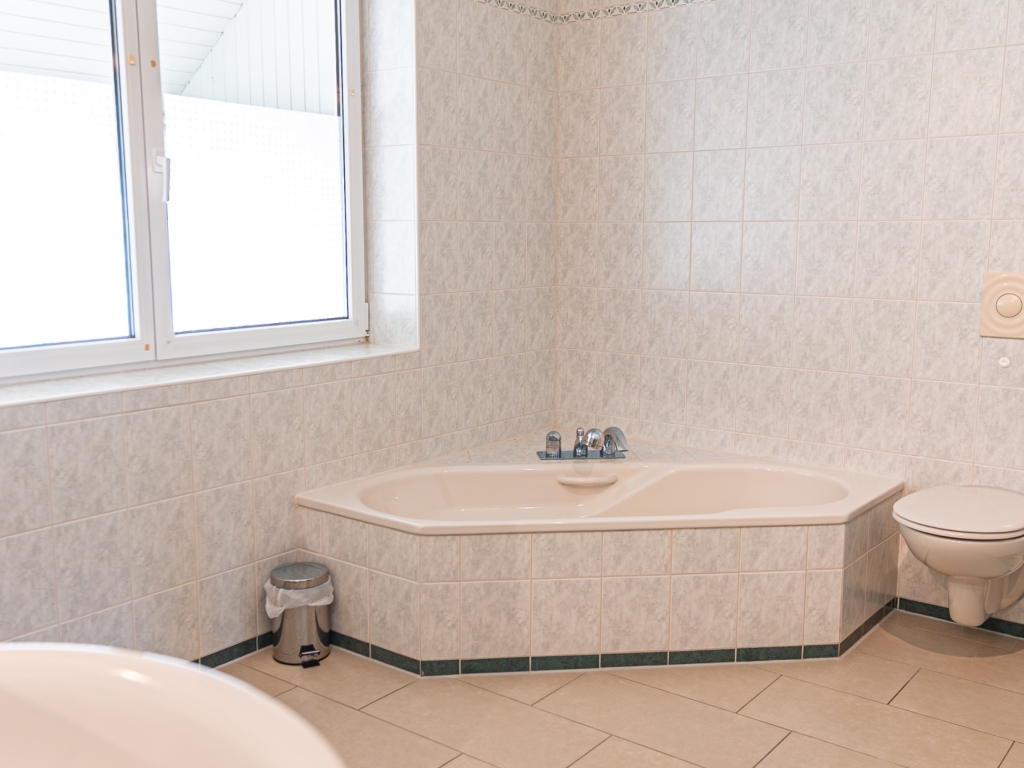 Ferienwohnung 8: Badezimmer mit Eckbadewanne, WC, Waschbecken und Fenster, heller Fliesenboden