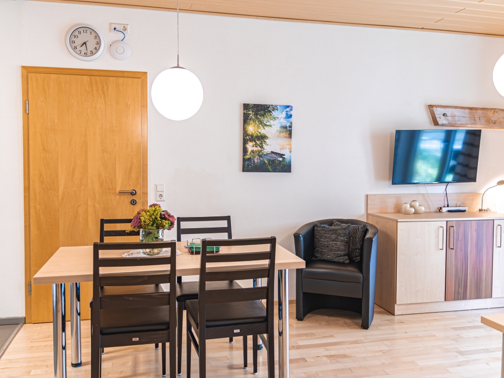 Ferienwohnung 8: Essbereich mit Holztisch und dunklen Stühlen, dahinter Sessel und Fernseher im Wohnraum