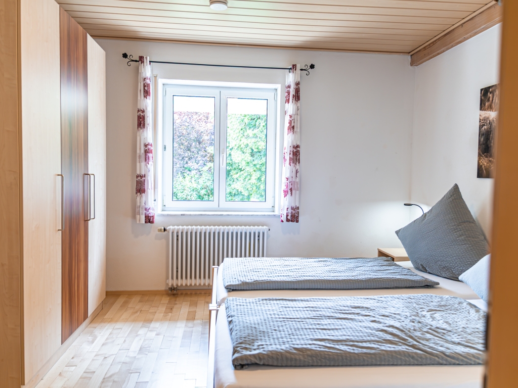 Ferienwohnung 9: Schlafzimmer mit Doppelbett, grauer Bettwäsche, Kleiderschrank und Fenster mit gemusterten Vorhängen
