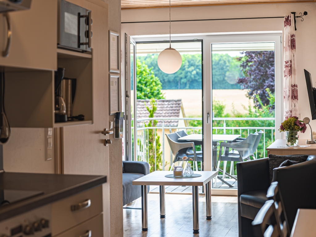 Ferienwohnung 9: Küchenzeile im Vordergrund, dahinter Couchtisch, Sessel und Balkontür mit Blick ins Grüne