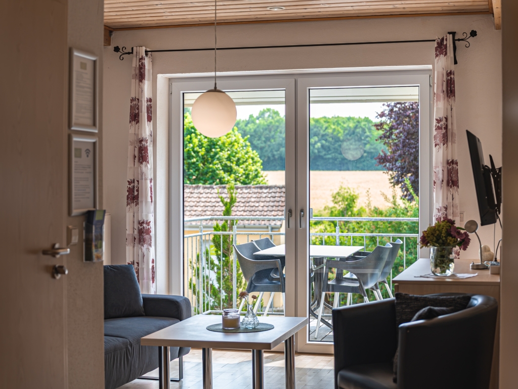 Ferienwohnung 9: Wohnzimmer mit Sesseln und Couchtisch, Balkontür mit gestreiften Vorhängen und grünem Ausblick
